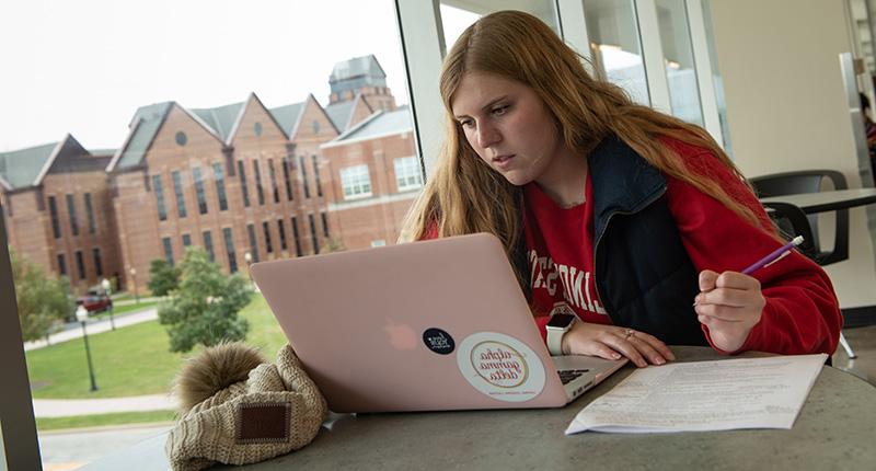 Female in front of a laptop at a table with the State Farm Hall of Business building in the background.